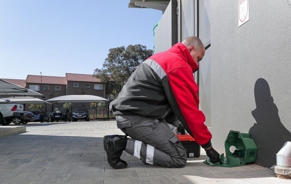 A Rentokil technician maintaining a green rodent bait station outside a commercial building.