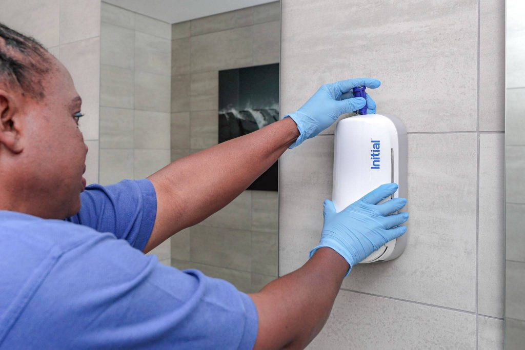 An Initial technician in gloves performs a professional service on a wall-mounted washroom dispenser.