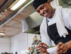 A smiling chef in a white uniform and black apron preparing food in a commercial kitchen