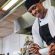 A smiling chef in a white uniform and black apron preparing food in a commercial kitchen