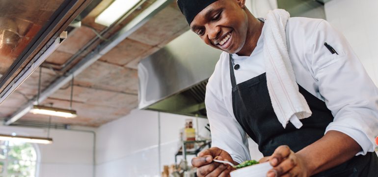 A smiling chef in a white uniform and black apron preparing food in a commercial kitchen